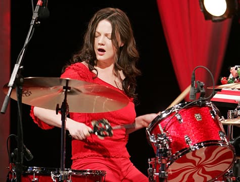 Meg White spielt ein rotes Schlagzeug auf einer Bühne mit roten Vorhängen im Hintergrund.