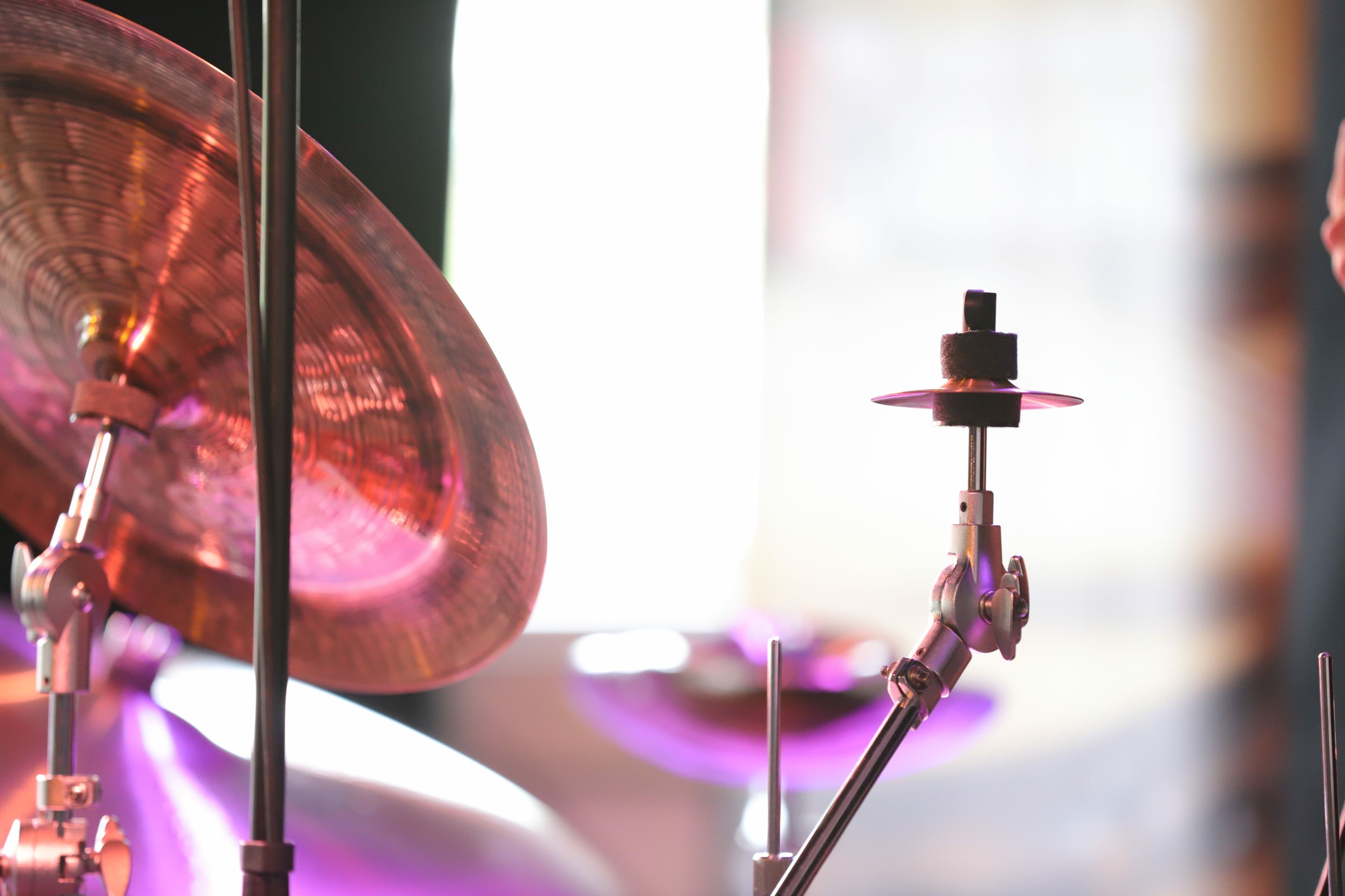 Nahaufnahme eines bronzefarbenen Schlagzeugbeckens und Metallständers, beleuchtet durch atmosphärisches pinkes und violettes Bühnenlicht.